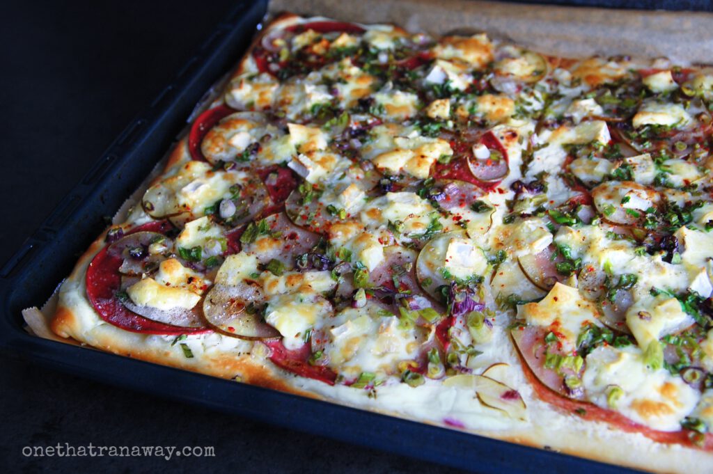 a black baking tray with baked dough, slices of salami, brie, pear and scallions on top