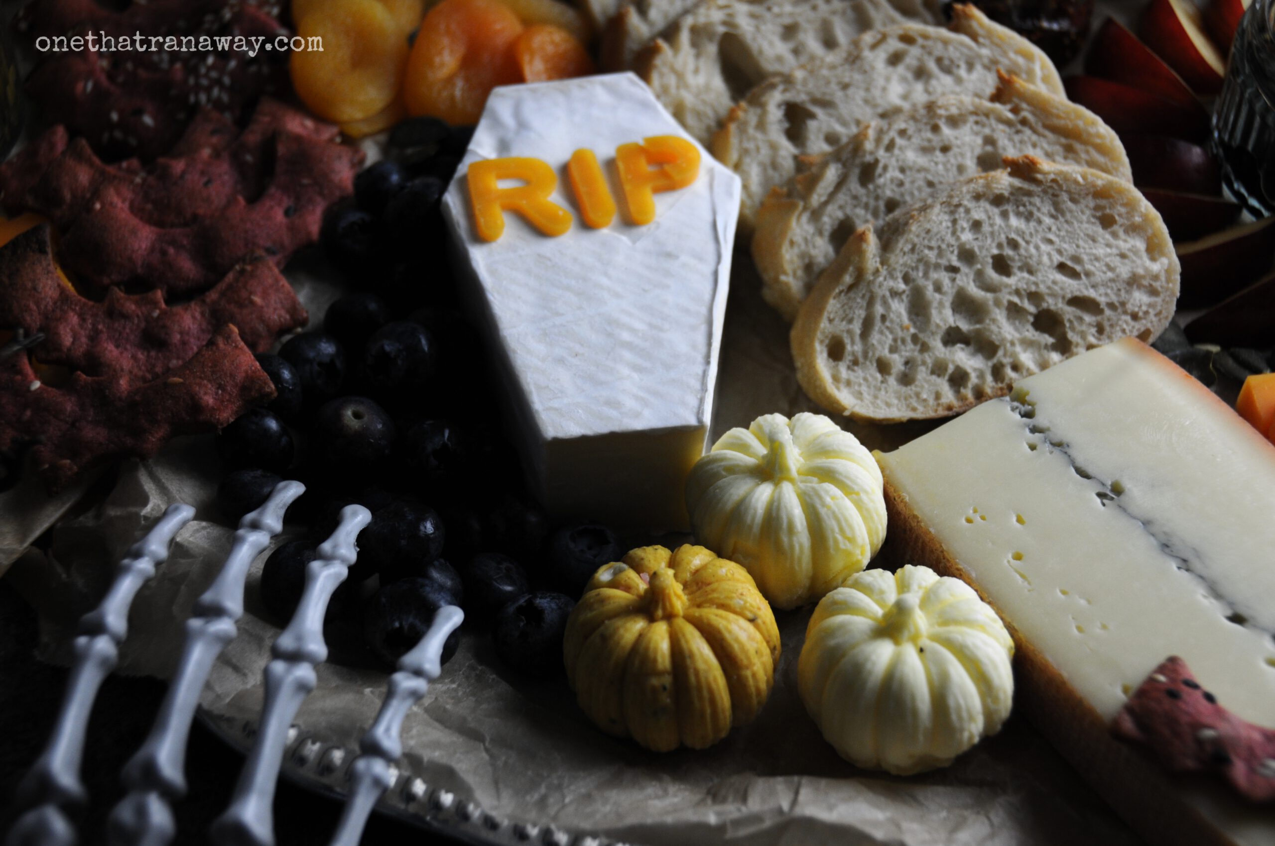 a Halloween themed cheese platter with a coffin shaped brie cheese, butter shaped like pumpkins and a fake skeleton hand