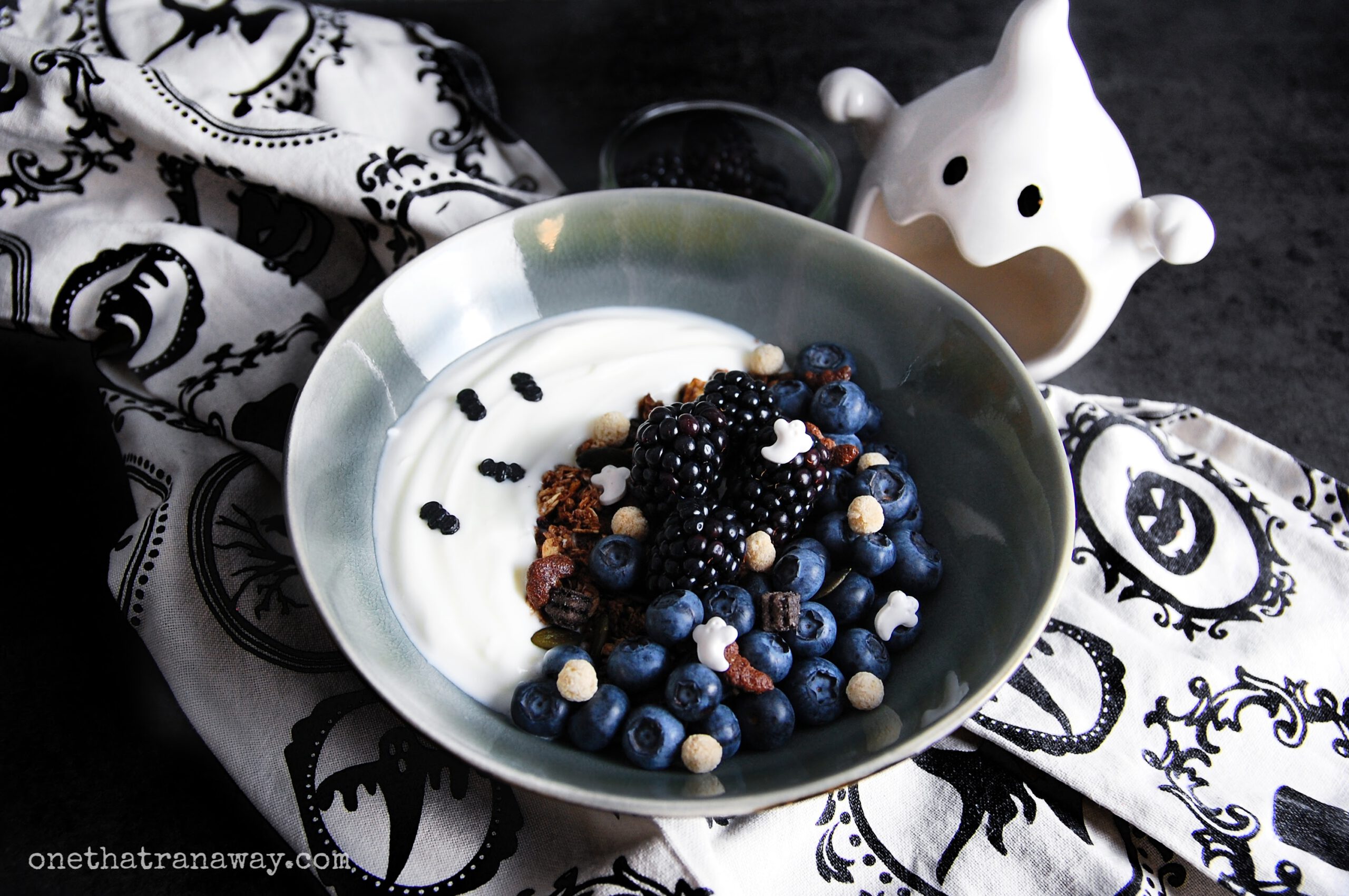 a bowl of yoghurt and cereal forming a crescent moon on top of a beige and black kitchen towel with a ceramic ghost in the background