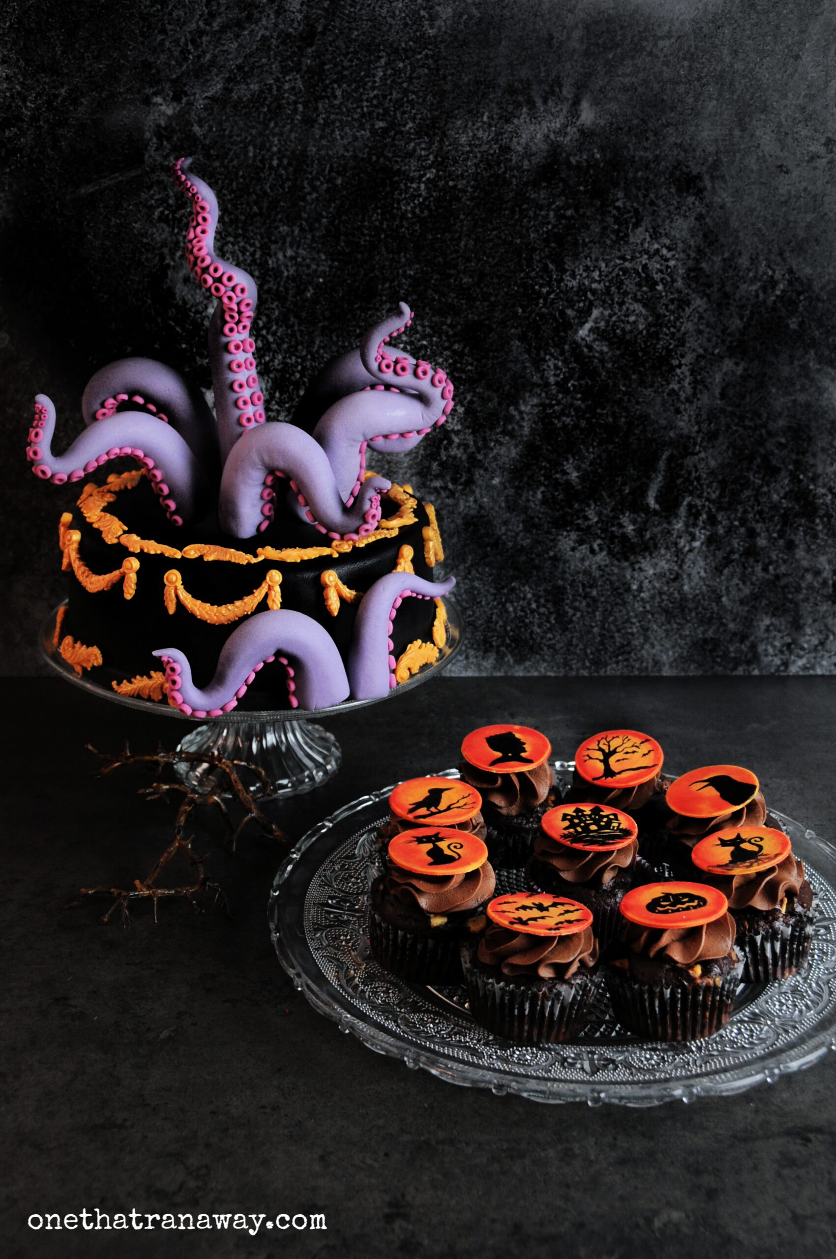 chocolate cupcakes with orange fondant toppers showing Halloween themed silhouettes on black background and a cake with fondant tentacles next to it