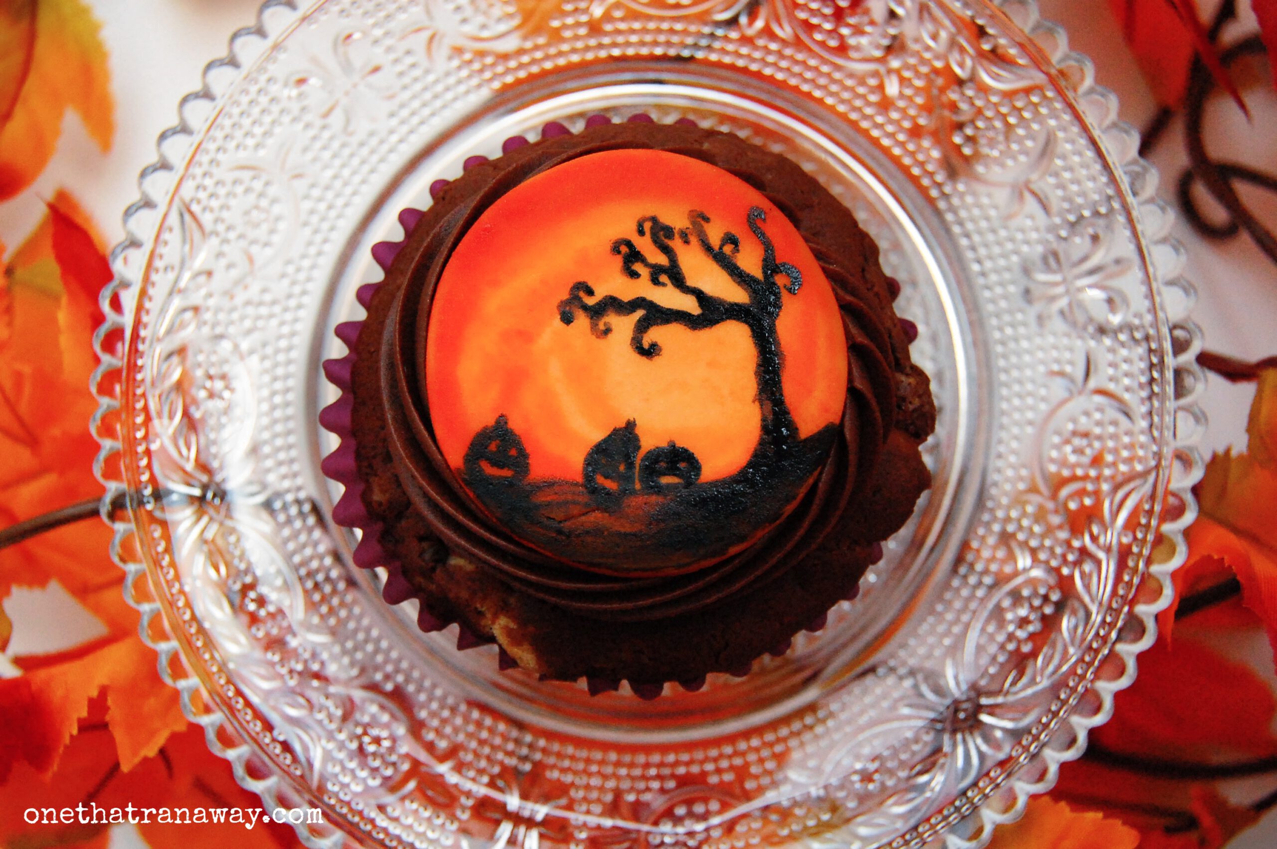 chocolate cupcake with an orange fondant topper showing the silhouette of a tree and pumpkins
